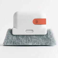 A white multi-surface gap cleaning brush with an orange scouring pad resting on a gray textured surface.