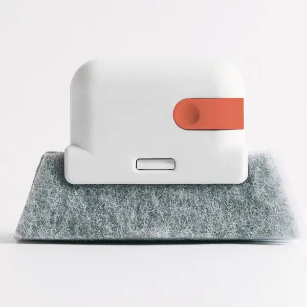 A white multi-surface gap cleaning brush with an orange scouring pad resting on a gray textured surface.
