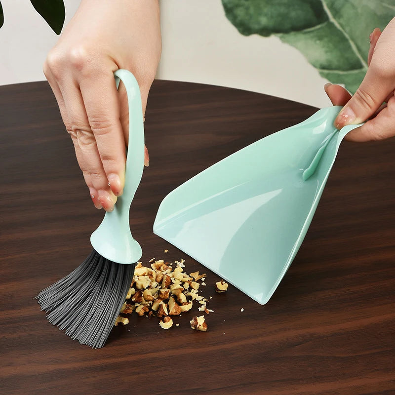 A hand is using a compact mini broom and dustpan set to clean a dark wooden surface.