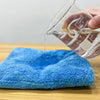 A hand pours liquid into a clear measuring cup, dripping onto a bright blue microfiber cleaning cloth on a wooden surface.