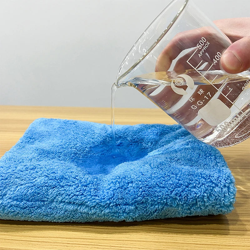 A hand pours liquid into a clear measuring cup, dripping onto a bright blue microfiber cleaning cloth on a wooden surface.