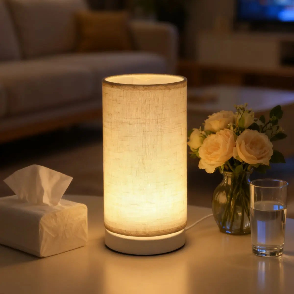 A modern white table lamp with a fabric shade emits a warm glow on a wooden surface.