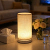 A modern white table lamp with a fabric shade emitting a warm glow on a light-colored surface.