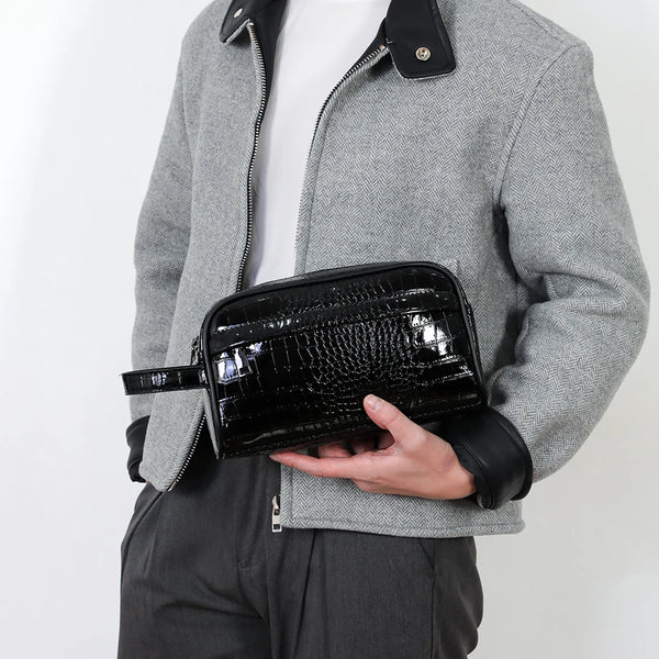 A man wearing a gray jacket holds a black crocodile-skin leather clutch bag with a detachable strap.