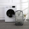 A white foldable laundry hamper with a mesh design sits on a gray carpet next to a washing machine.