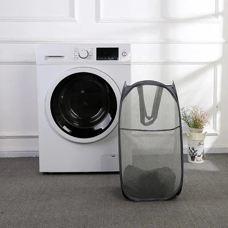 A white foldable laundry hamper with a mesh design sits on a gray carpet next to a washing machine.