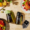 A dark green glass bottle of extra virgin olive oil dispenser and sprayer next to a lemon and a plate of roasted poultry.