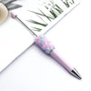 A pink ballpoint pen with a sparkly diamond sequin filling is shown next to green foliage.