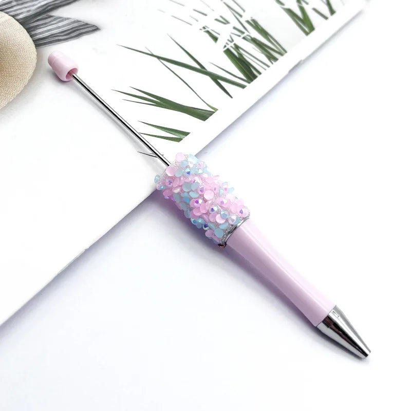A pink ballpoint pen with a sparkly diamond sequin filling is shown next to green foliage.