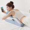 A woman is lying down on a white bed using a light blue memory foam full-body support pillow while looking at her smartphone.