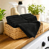 A stack of black microfiber cleaning towels is displayed in a woven basket on a wooden surface.