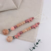 A string of silicone pacifier clips with baby names and pink bows displayed on a beige linen fabric.