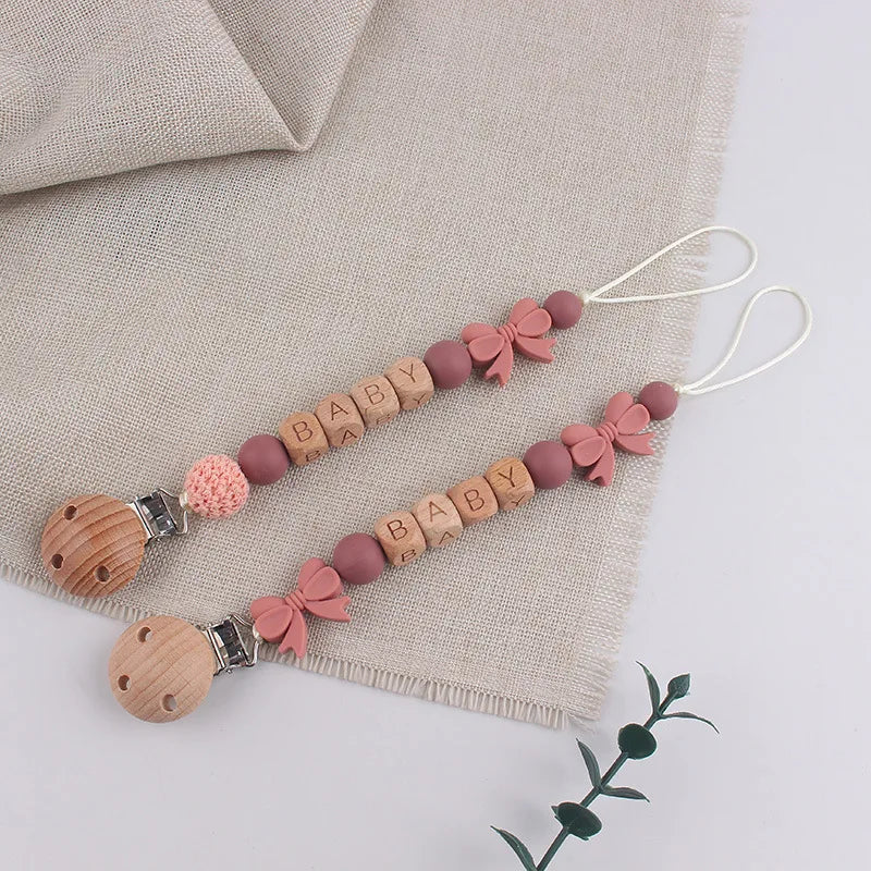 A string of silicone pacifier clips with baby names and pink bows displayed on a beige linen fabric.