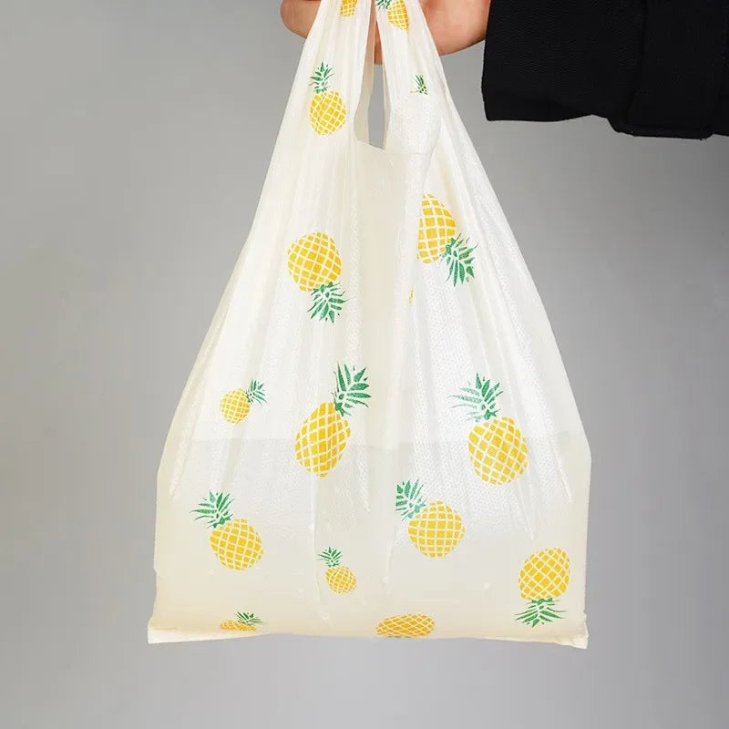 A white mesh tote bag with yellow pineapple print and handles is shown against a gray background.