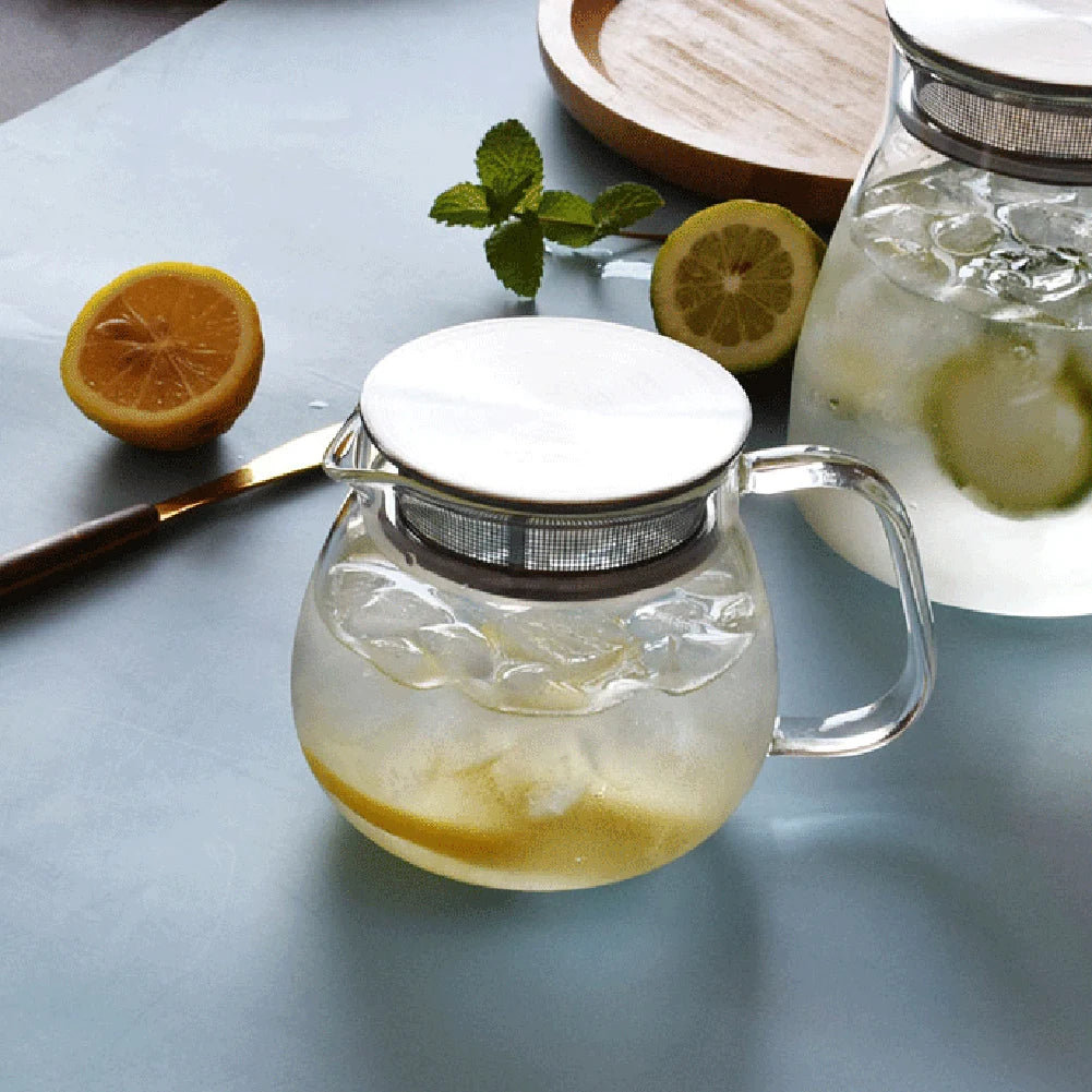 A clear glass teapot with a stainless steel filter filled with ice and lemon slices on a light blue surface.