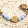 A wooden pacifier clip with a chain of light blue and beige crochet beads resting on a cream-colored knitted surface.
