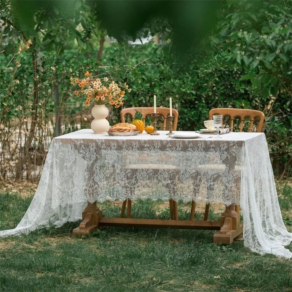 An elegant white lace tablecloth set for a table outdoors with greenery and floral accents.