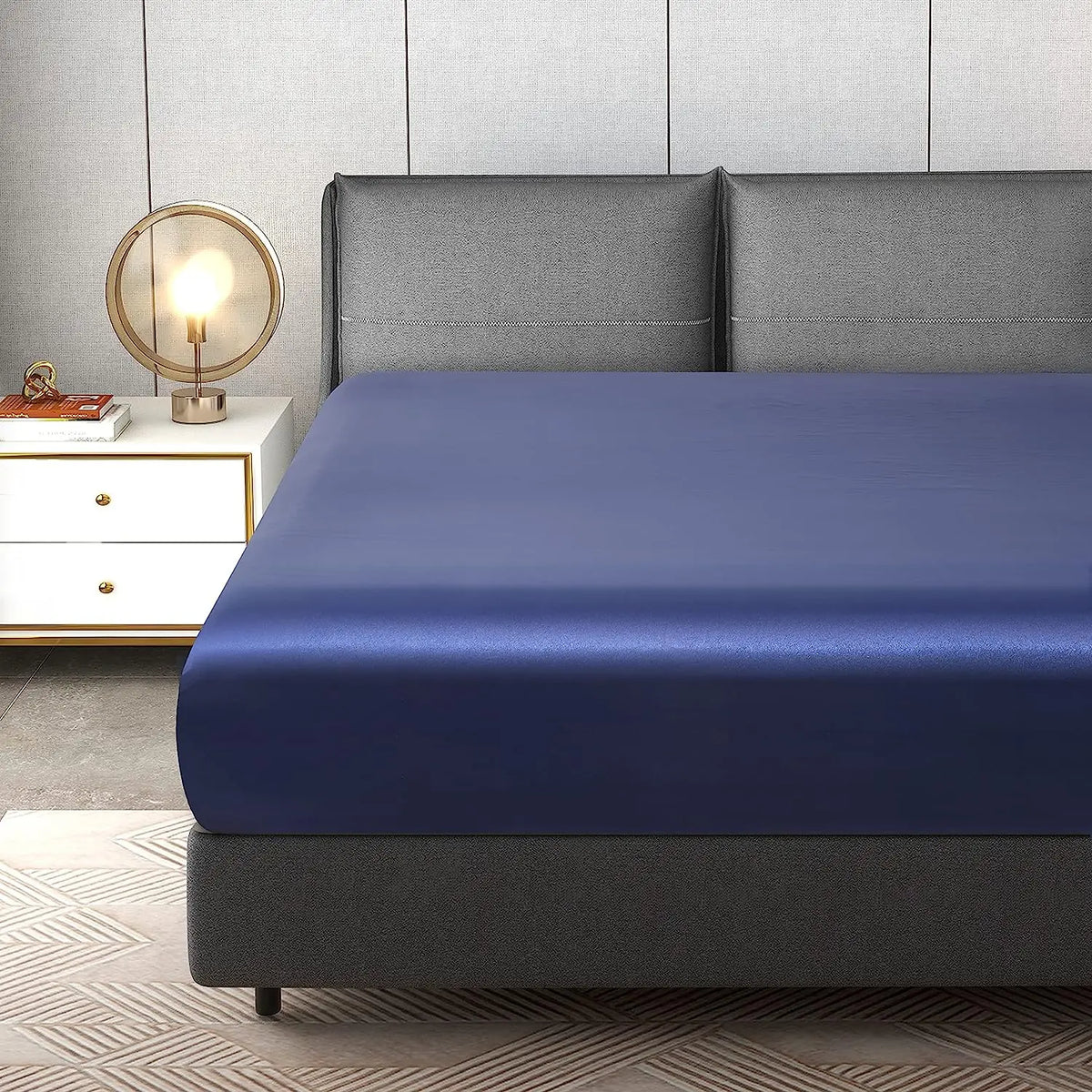 A navy blue luxury satin fitted sheet is displayed on a bed with a gray headboard and bedside lamp.