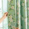 A close-up of a light green curtain panel featuring a white and pink floral plant pattern, being held by a hand next to a win