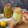 Hands are squeezing a glass mason jar filled with sliced cucumbers and herbs on a wooden cutting board.