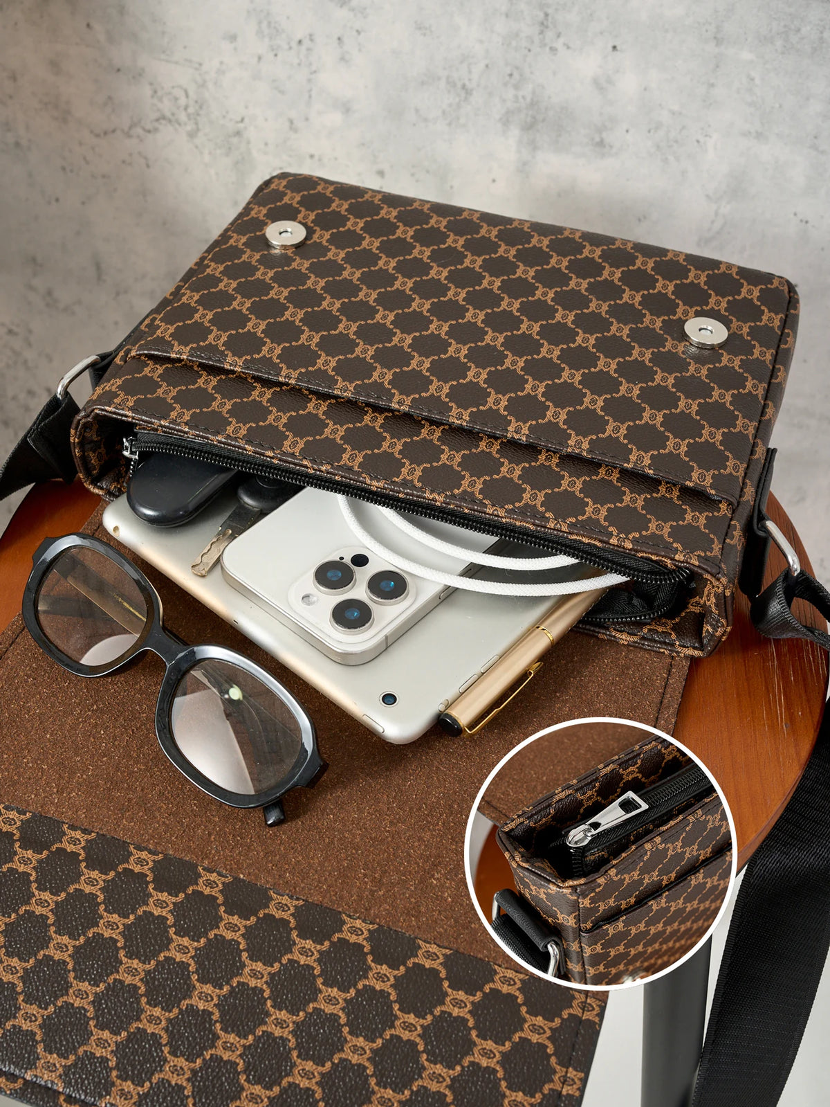 A brown Gucci print crossbody bag is open, revealing a smartphone and sunglasses on a wooden surface.