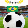 A white and black realistic football plush pillow is centered on green grass with a Brazilian flag in the background.