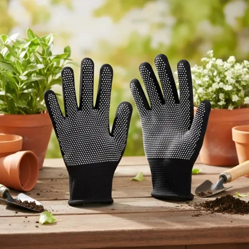 A pair of black knitted work gloves with a dotted grip are displayed on a wooden surface with potted plants in the background