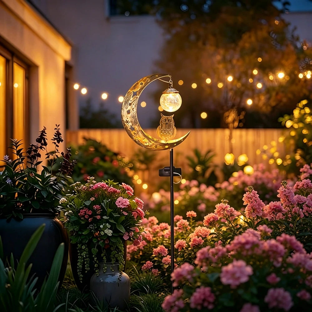 A solar owl garden light with a crescent moon decoration illuminates pink flowers in a garden setting.