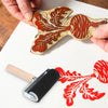 A hand is pressing a rubber ink roller onto paper, creating a red leaf print on the surface.