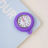 A purple silicone pocket watch with a silver face and crown is shown resting on a light blue notebook.