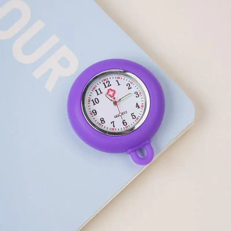 A purple silicone pocket watch with a silver face and crown is shown resting on a light blue notebook.