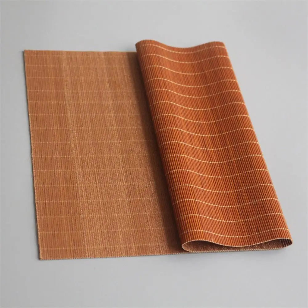 A rectangular, woven bamboo tea mat in a warm orange-brown color is shown rolled up next to its flat form.
