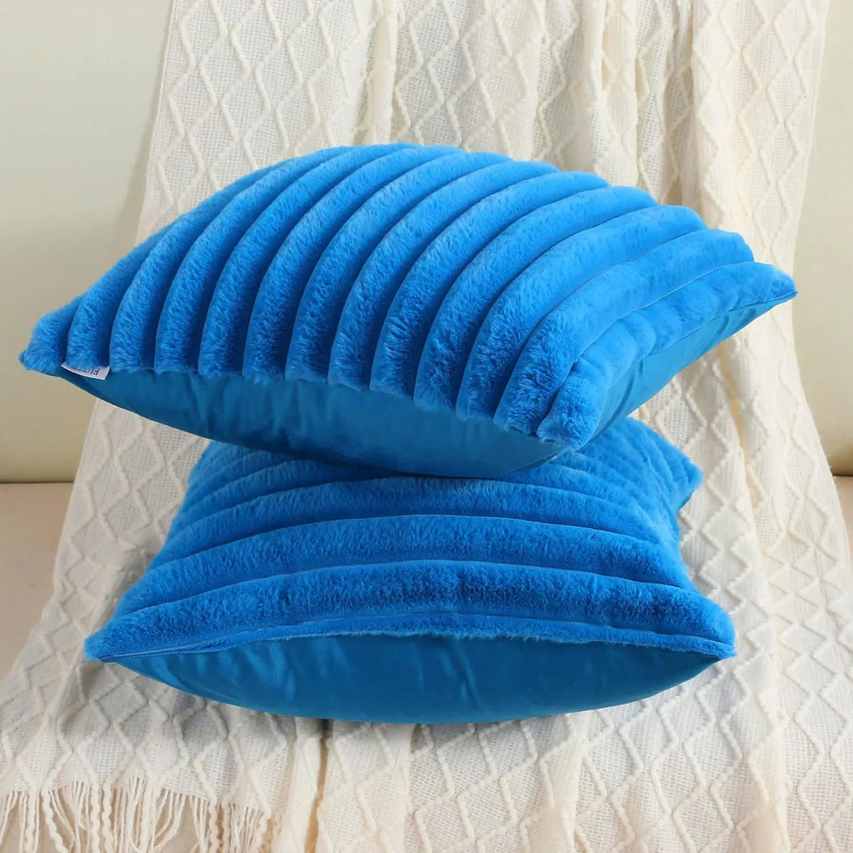 A bright blue plush faux fur square cushion cover with a ribbed stripe detail resting on a white textured fabric.
