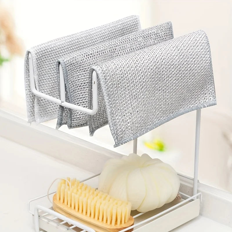 A set of ten reusable steel wire scrubber dishcloths hanging on a white rack next to a yellow brush and a white squash.