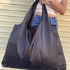 A person is holding a large black foldable nylon shopping tote bag against a light-colored background.