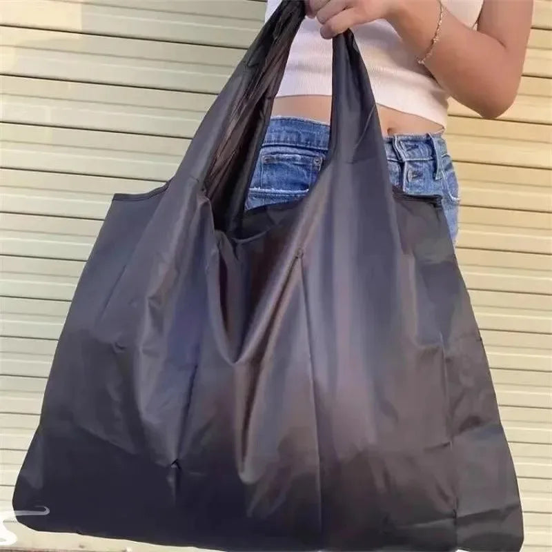 A person is holding a large black foldable nylon shopping tote bag against a light-colored background.