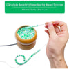 A hand is using clip-style beading needles for a bead spinner, showing the needles and a bowl of green beads.