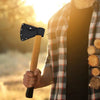 A hand holds a wooden handle with a black nylon axe blade cover and belt loop against a bright, outdoor background.