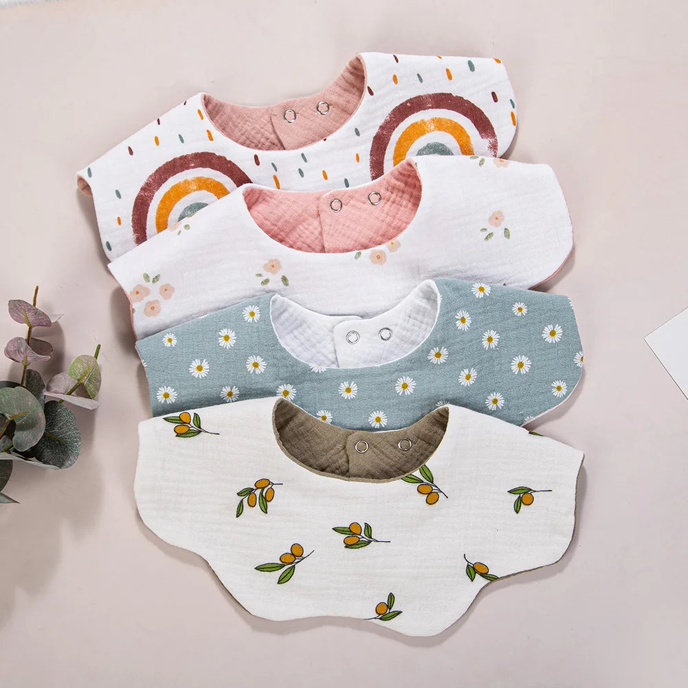 A collection of five white muslin cotton baby bibs and burp cloths featuring various patterns including rainbows, florals, an