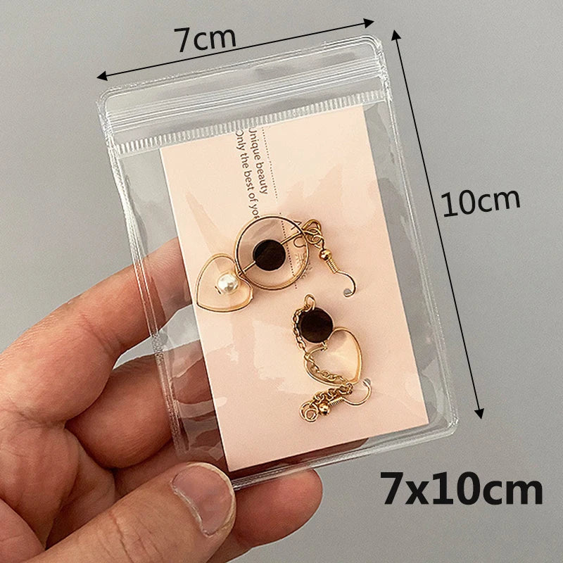 A hand holds a clear plastic pouch containing two gold-toned jewelry pieces, including a hoop earring and a pendant.