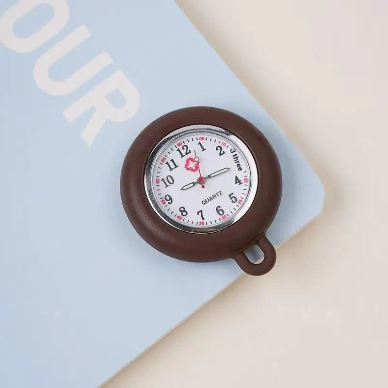 A brown silicone pocket watch with a clear face and a pink floral detail is shown resting on a light blue notebook.