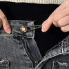 A hand is adjusting an adjustable metal button on the waistband of dark denim jeans.