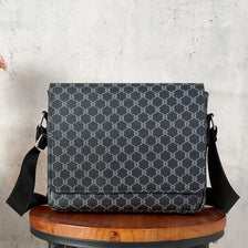 A dark gray patterned crossbody bag resting on a wooden stool against a light gray background.
