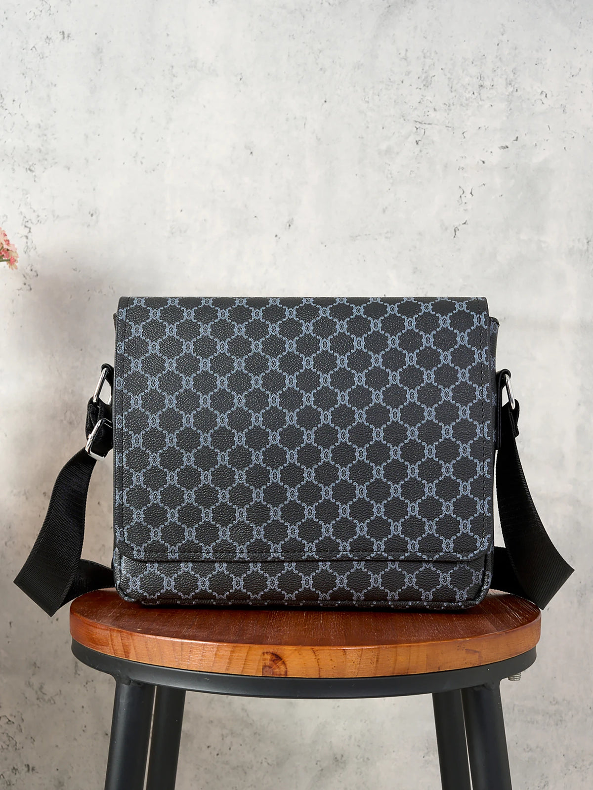 A dark gray patterned crossbody bag resting on a wooden stool against a light gray background.