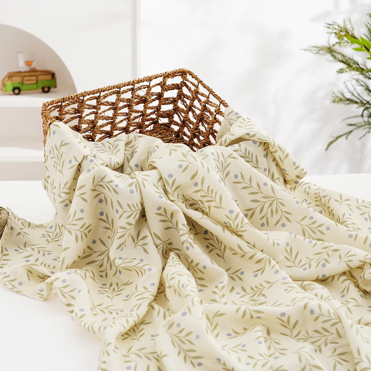 A soft, light beige muslin baby swaddle blanket with a delicate green leaf pattern is draped on a white surface.