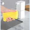A hand is using a yellow sponge to clean a white touchless soap dispenser on a kitchen counter.