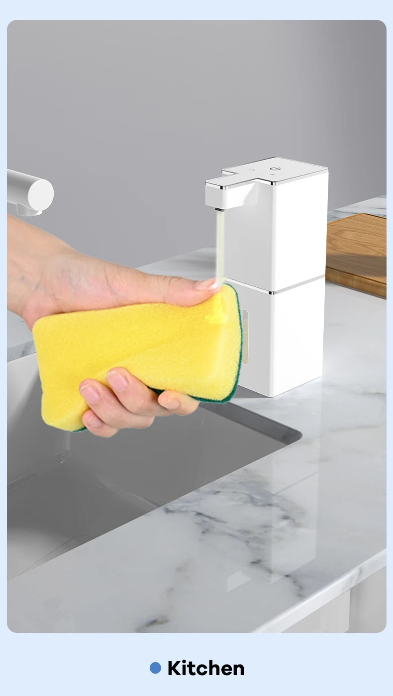 A hand is using a yellow sponge to clean a white touchless soap dispenser on a kitchen counter.