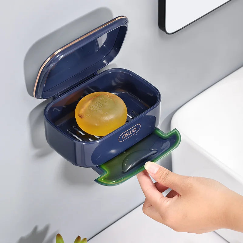 A hand is opening a blue two-tier draining soap holder with a yellow soap ball in a bathroom setting.