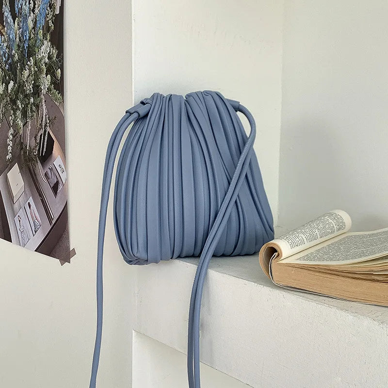 A light blue pleated faux leather crossbody bag hanging on a white shelf next to a stack of books.