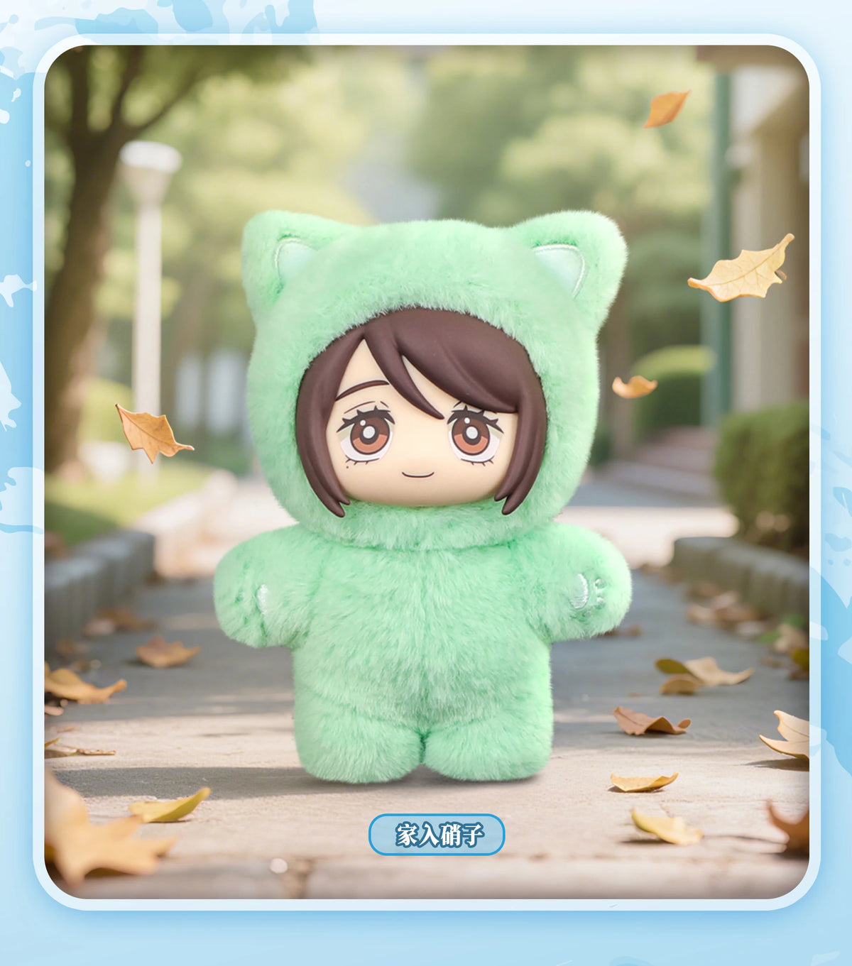 A soft green cat plush doll pendant in a hoodie is displayed outdoors on a wooden path with autumn leaves.